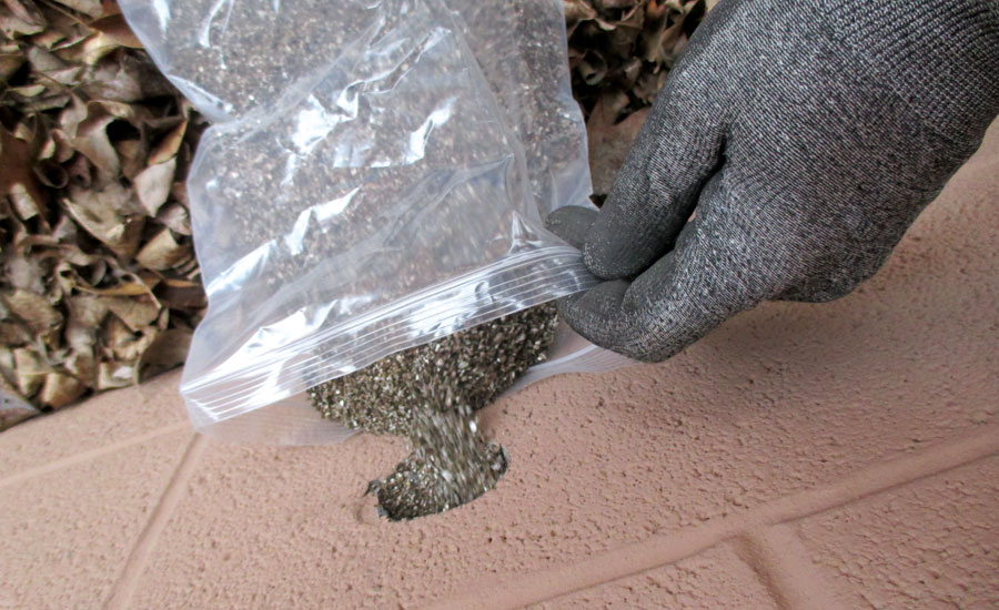 A gloved hand holding a plastic bag to a wall that is pouring out mercury which is a grainy silver substance. Reusing building material is prohibited for mercury